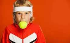 boy in tennis gear with tennis ball in his mouth and angry expression