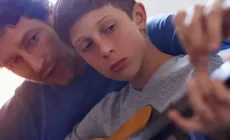 dad leans over son's shoulder to teach guitar