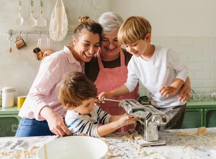 Une famille avec deux jeunes garçons qui font des pâtes ensemble dans la cuisine, guidés par leur mère et leur grand-mère.